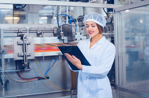 A young beautiful girl in white overalls makes notes in a tablet on the background of equipment of a food processing plant. Quality control in production