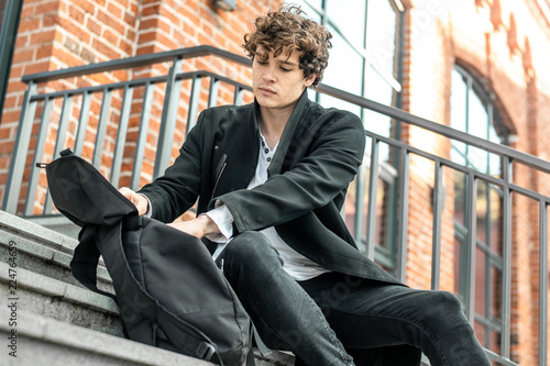 Tableau sur toile Young attractive pensive man sitting on steps and rummaging in his backpack