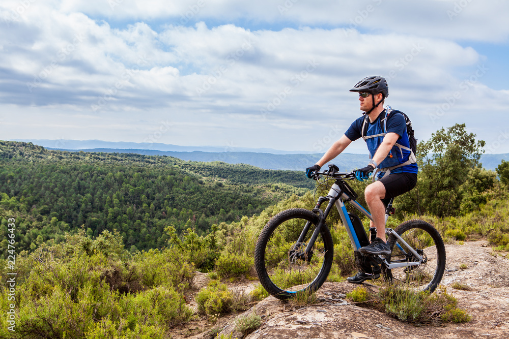 Mountain biker wearing sunglasses looking into distance on top of hill, holding one foot on the pedal