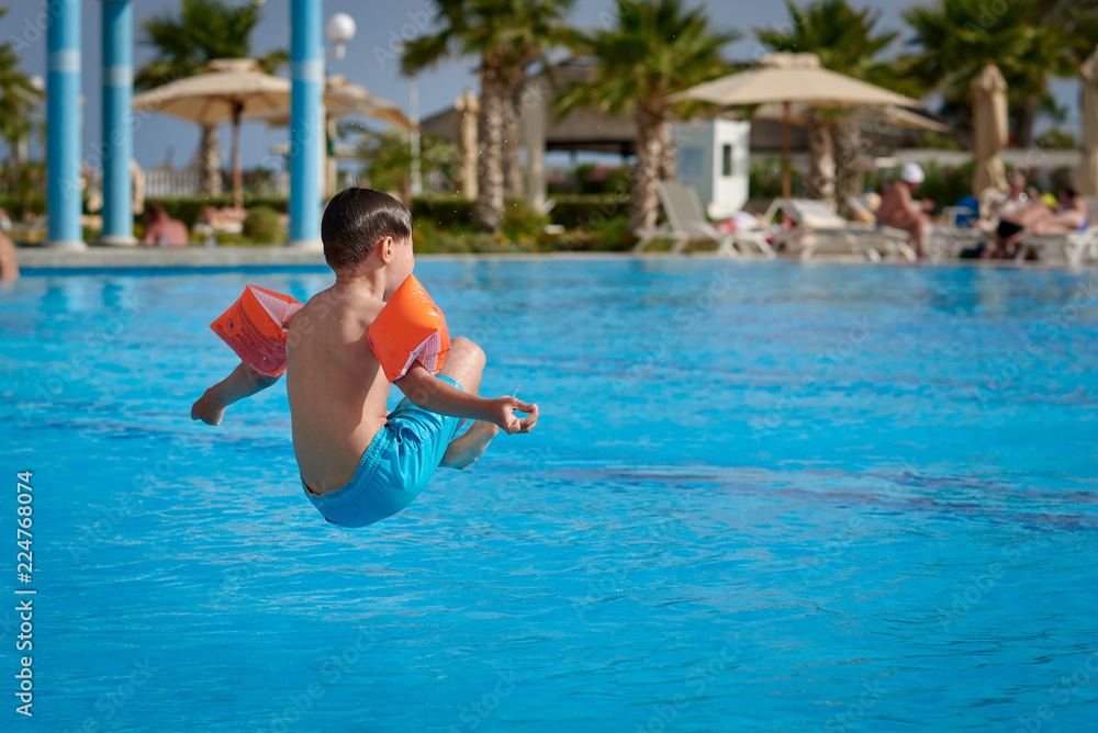 Caucasian boy having fun making fantastic jump into swimming pool at resort. He has pose of sitting man. Side view.