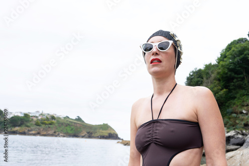 Portrait of a woman by the beach with a 1950's swim hat and sunglasses on posing on a rock