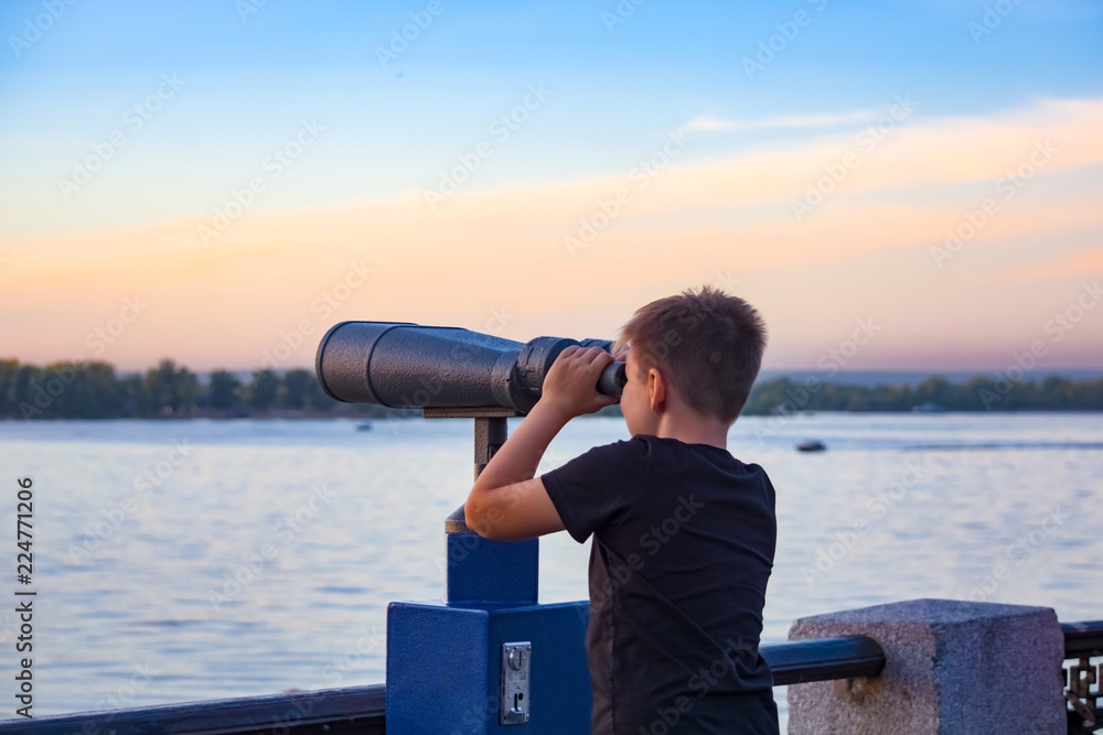 Obraz premium The boy looking through binoculars by the river at sunset