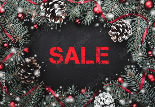 Black stone background with a frame of fir branches decorated with balls and red ribbon. Pine cones. Top view. Red text sale.