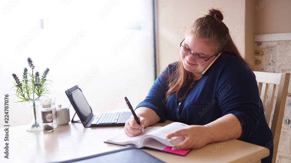 Plus size business woman busy multitasking at her local coffee shop to get as much work done as possible early in the morning, as she writes in her journal and talks on the phone simultaneously. Plus size business woman busy multitasking at her local coffee shop to get as much work done as possible early in the morning, as she writes in her journal and talks on the phone simultaneously.