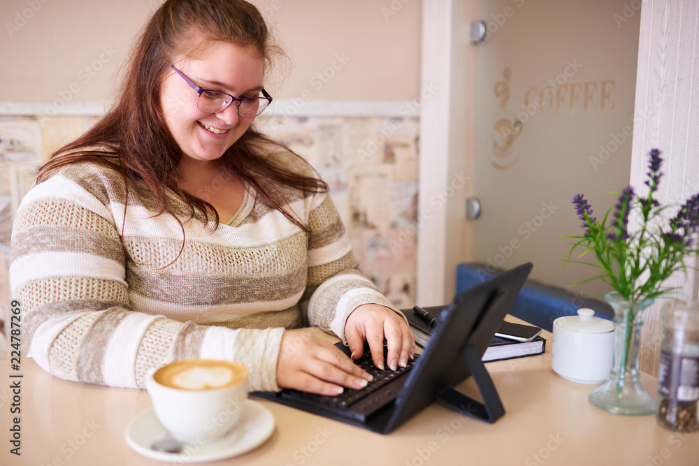 Plus size caucsian woman happily smiling while working on her tablet and keyboard in a bright cafe with a morning cup of coffee to be more productive. Plus size caucsian woman happily smiling while working on her tablet and keyboard in a bright cafe with a morning cup of coffee to be more productive.