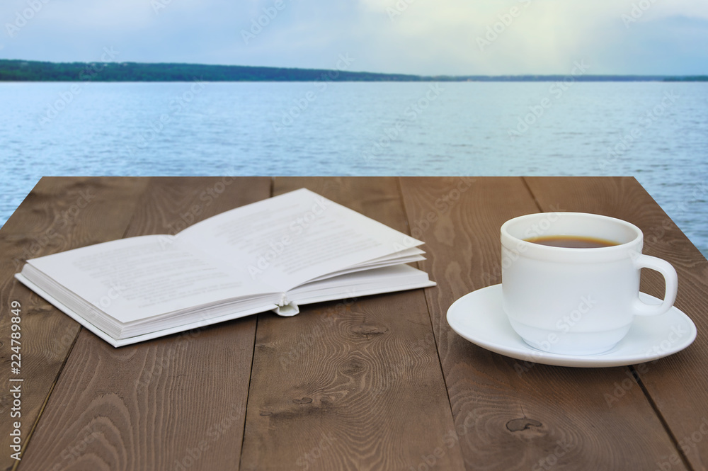 White cup with coffee on a saucer and a book. On a wooden brown background. View of the sea.