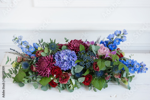 Elongated floral arrangement in vintage metal vase. table setting. Blue and red color. Gorgeous bouquet of different flowers. Bright room, stucco on the wall