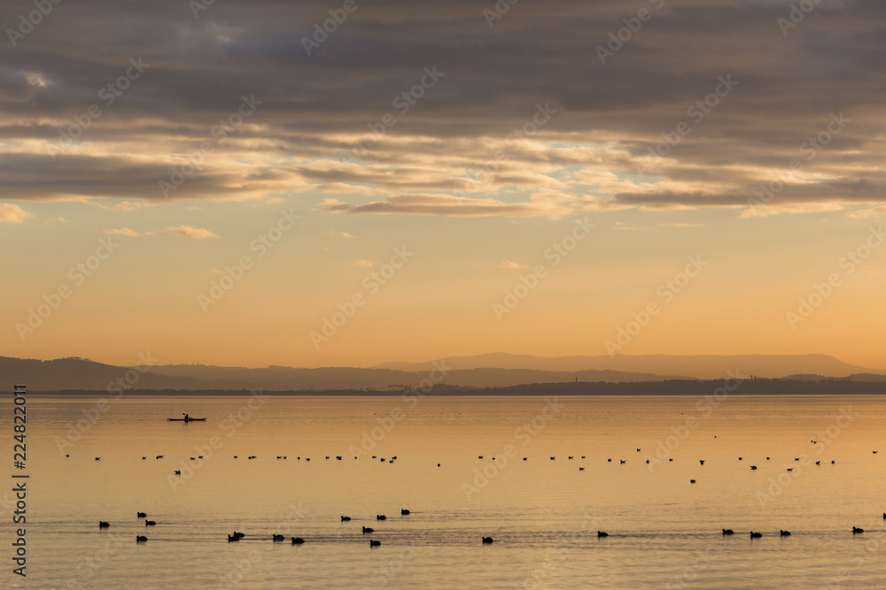 Beautiful view of a lake at sunset, with orange tones, birds on water and a man on a canoe Beautiful view of a lake at sunset, with orange tones, birds on water and a man on a canoe