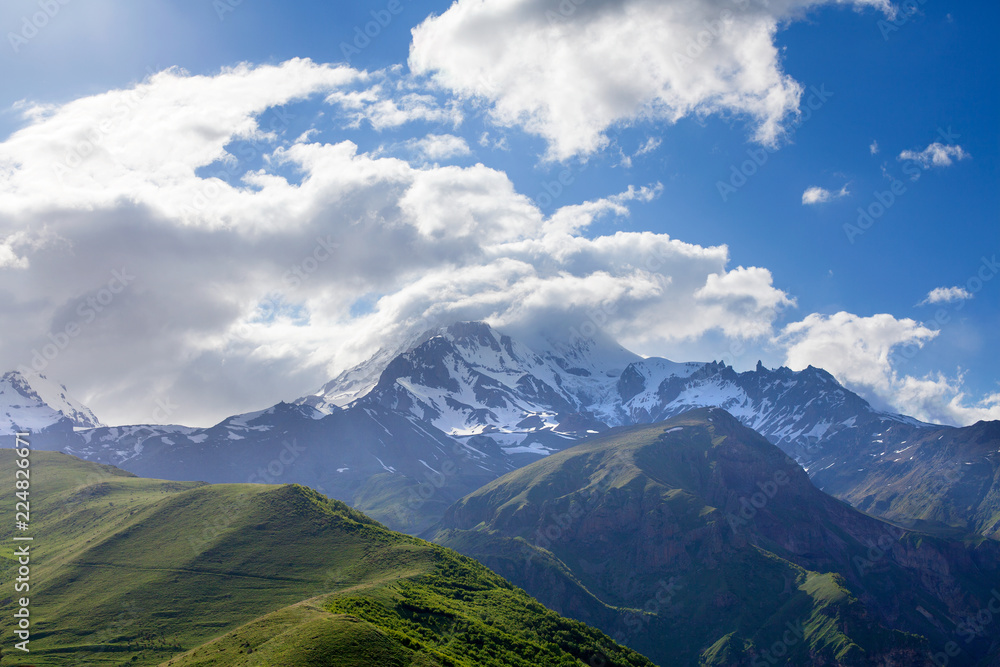 Panorama landscape mountain peaks in snow and green hills, deep blue sky and huge white clouds background, Caucasian mountains, Kazbek mountain, Georgia