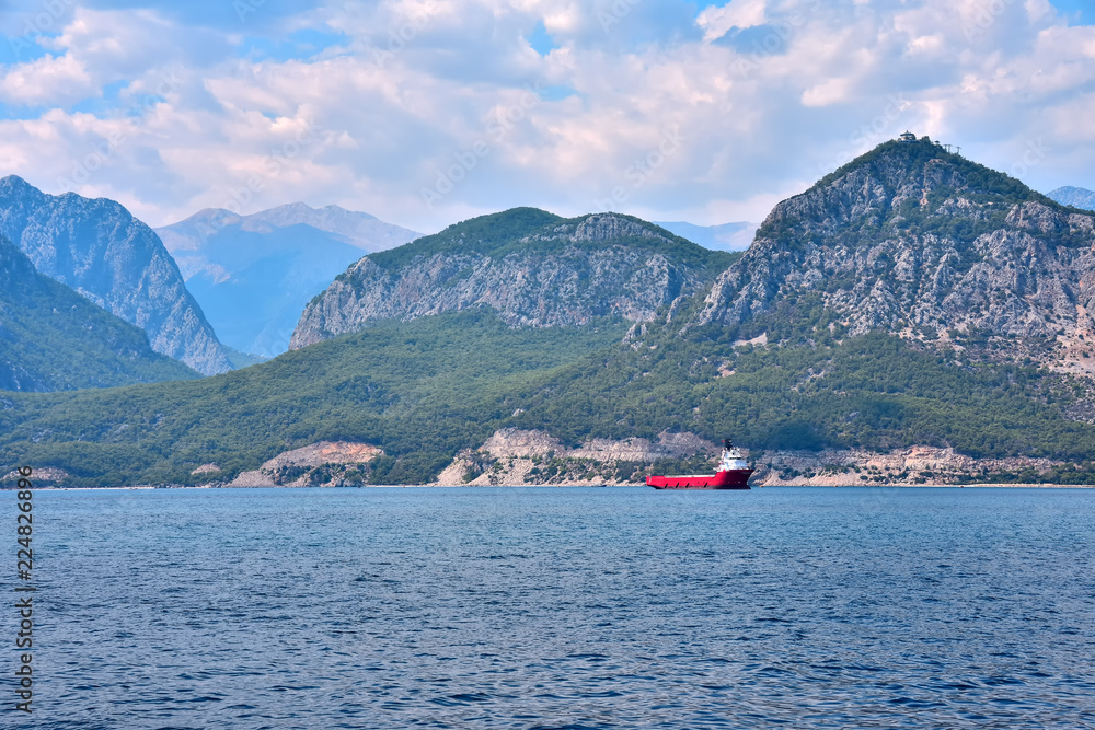 vessel for transportation of containers at the anchorage near the sea shore, against the background of mountains and sky covered with clouds