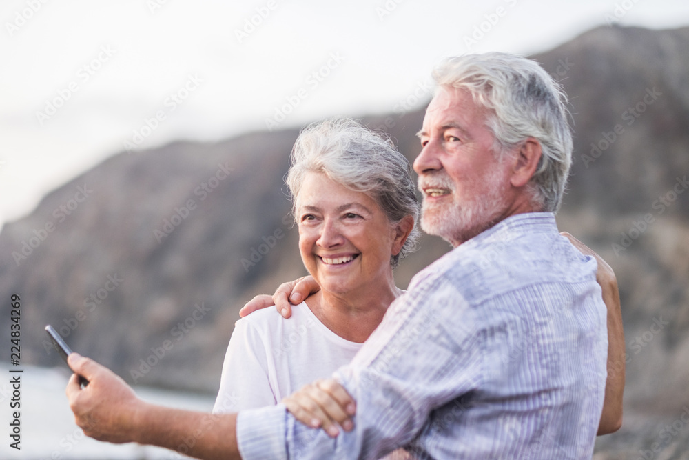 concept of vacation, technology, tourism, travel and people - happy senior couple with cell phone phone on pebble beach laughing and joking hugging and making photo. White hair