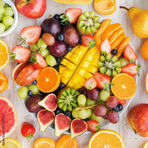 Variety of cut fruits and berries platter, strawberries blueberries, mango orange, apple, grapes, kiwis on the white wood background, copy space for text, square, top view, selective focus