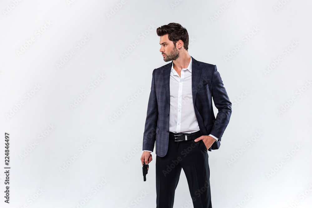Dangerous style. Handsome young man looking away and holding a gun while standing against white background Dangerous style. Handsome young man looking away and holding a gun while standing against white background
