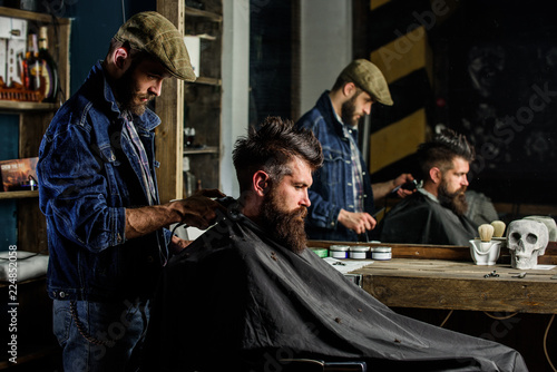 Barber styling hair of brutal bearded client with clipper. Hipster client getting haircut. Barber with hair clipper works on hairstyle for bearded man barbershop background. Hipster lifestyle concept