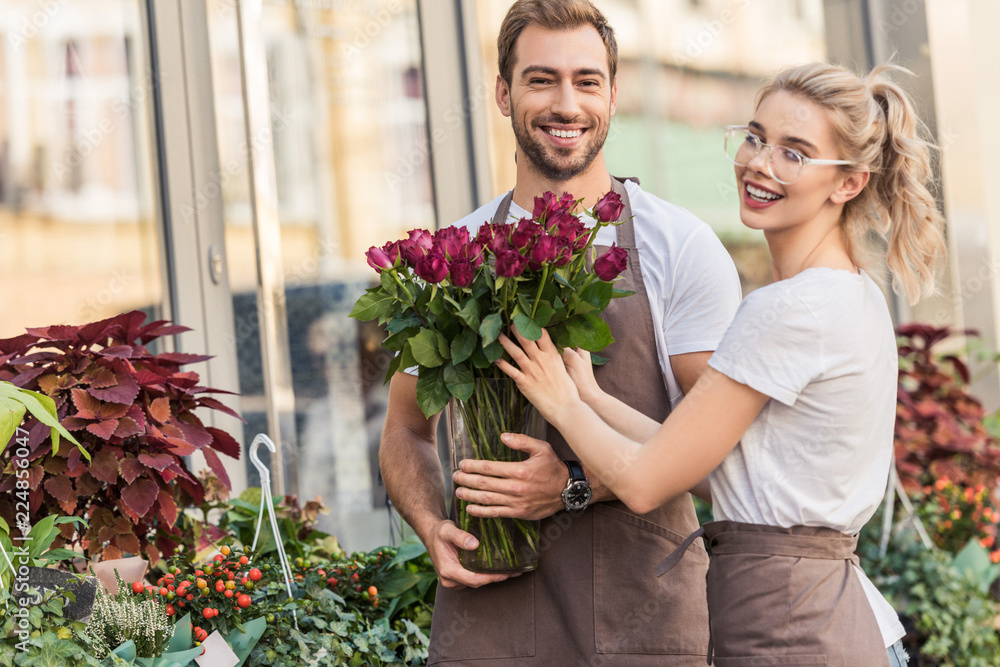 smiling florists holding burgundy roses near flower shop and looking at camera