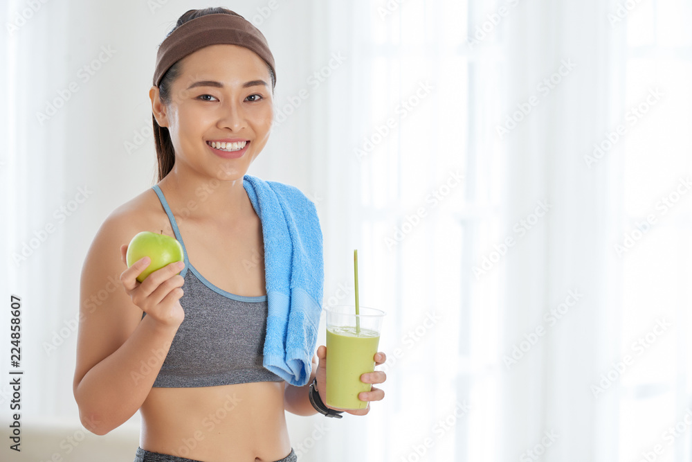 Happy Asian sportswoman holding ripe apple and green smoothie in cup smiling at camera Happy Asian sportswoman holding ripe apple and green smoothie in cup smiling at camera