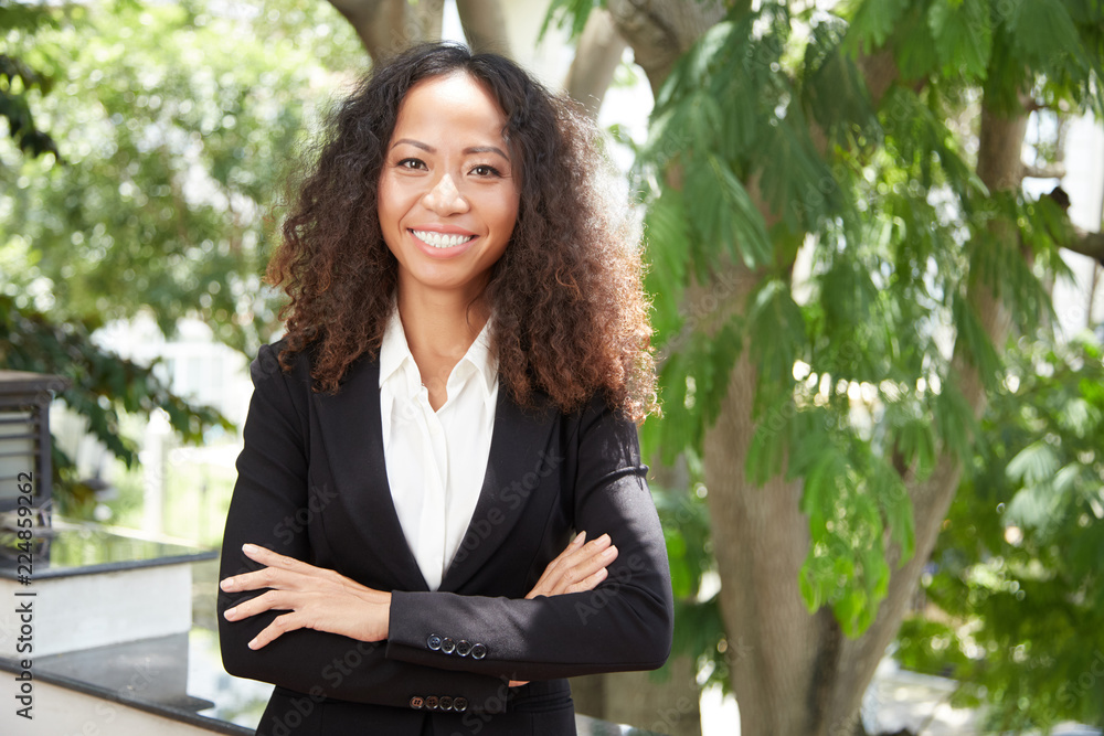 Young ethnic woman in formal outfit holding arm crossed and smiling at camera standing on balcony Young ethnic woman in formal outfit holding arm crossed and smiling at camera standing on balcony