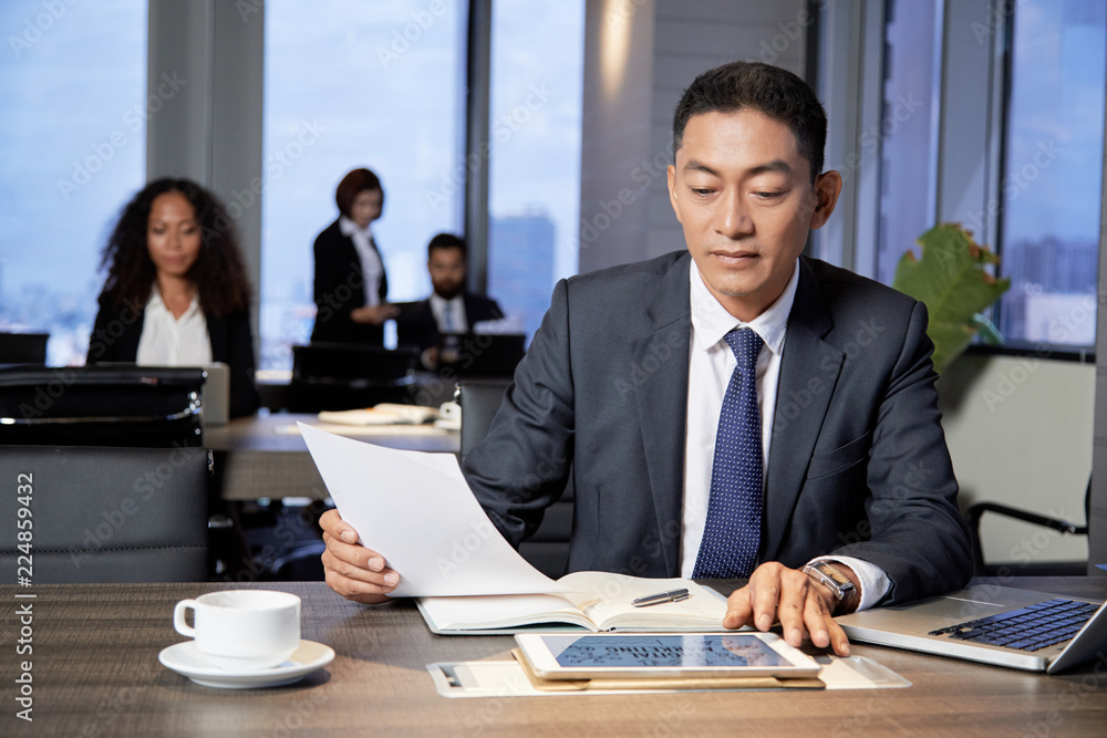 Adult Asian man with tablet and laptop sitting at table and reading papers in office with colleagues Adult Asian man with tablet and laptop sitting at table and reading papers in office with colleagues