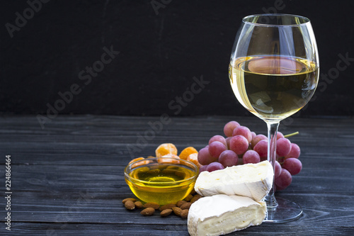 A glass of white wine with cheese cuts, figs, nuts, honey, grapes on a dark rustic wooden boards background
