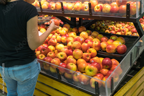 Fresh healthy fruits on shelves in supermarket, with female buyer choosing red apples