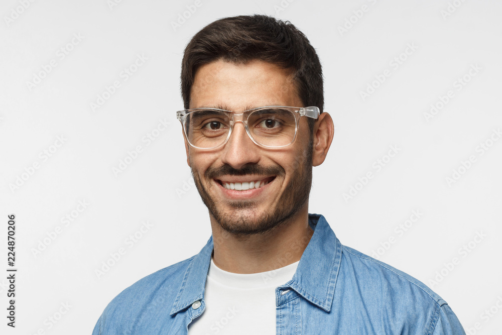 Closeup headshot of smiling young man in blue shirt against gray background, wearing transparent spectacles Closeup headshot of smiling young man in blue shirt against gray background, wearing transparent spectacles