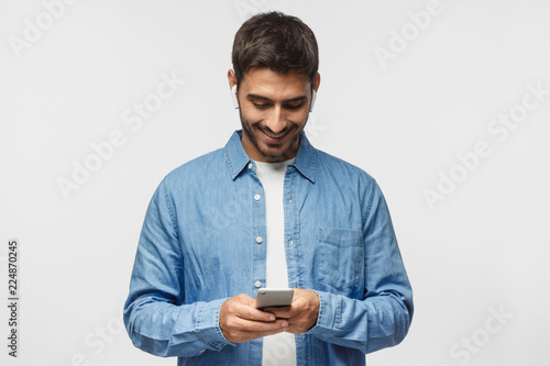 Young man standing isolated on gray background, looking at screen of smartphone, browsing web and smiling nicely while chatting © Damir Khabirov