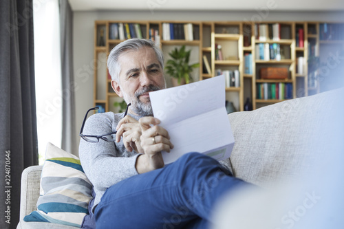 Mature man sitting on couch at home reading letter photo