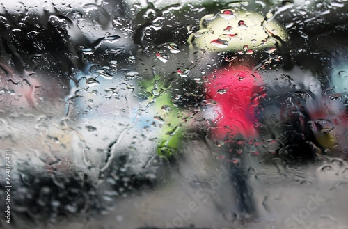 Abstract view through rain drops on a window pane of a woman in a red coat holding a yellow umbrella walking in the street past cars.