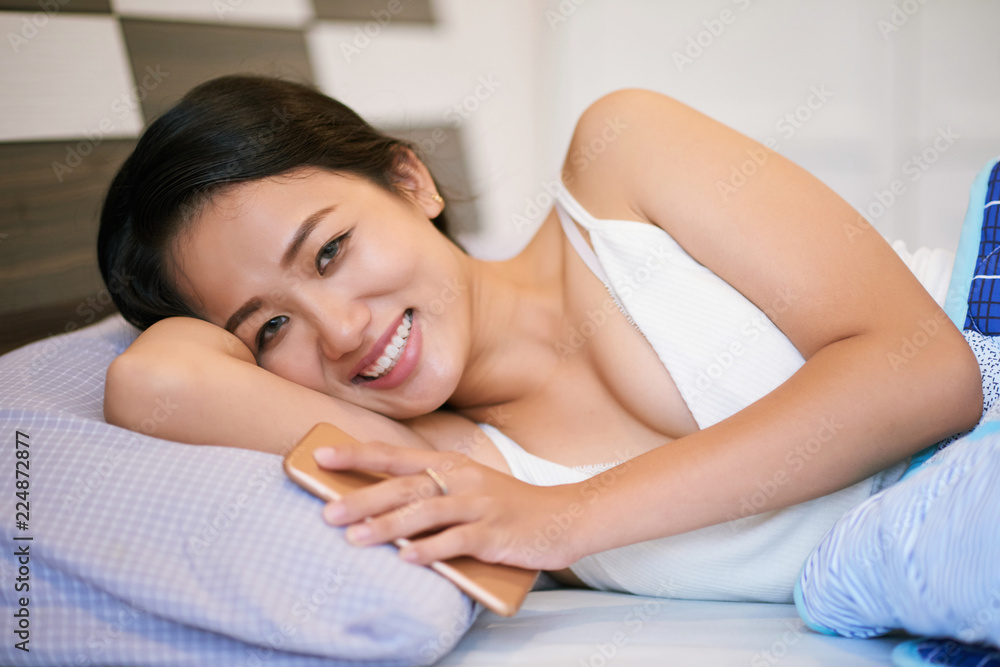 Young happy Asian woman lying in cozy bed holding smartphone and smiling charmingly at camera Young happy Asian woman lying in cozy bed holding smartphone and smiling charmingly at camera