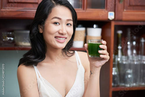 Young Asian woman in white top holding glass of green freshly made juice looking at camera at home
