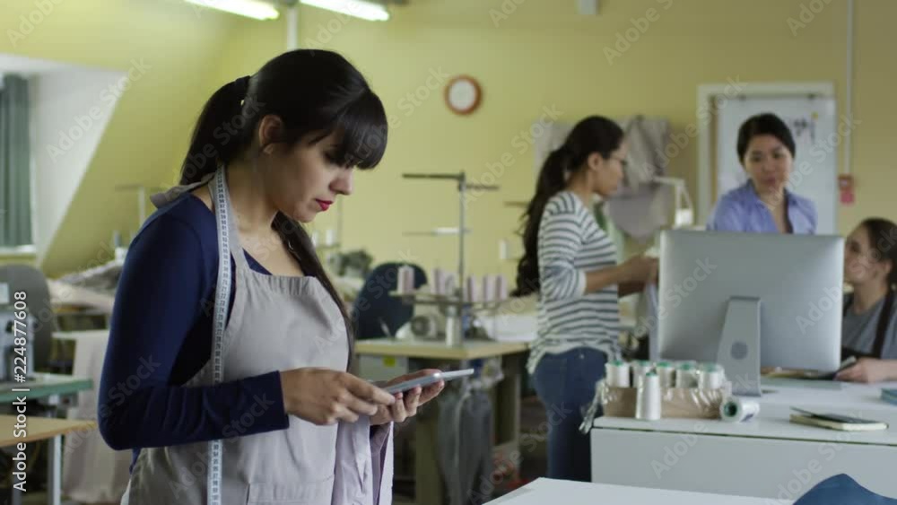 Medium shot of young Hispanic woman using tablet computer when working at sewing factory, her colleagues discussing fabric in the background Medium shot of young Hispanic woman using tablet computer when working at sewing factory, her colleagues discussing fabric in the background