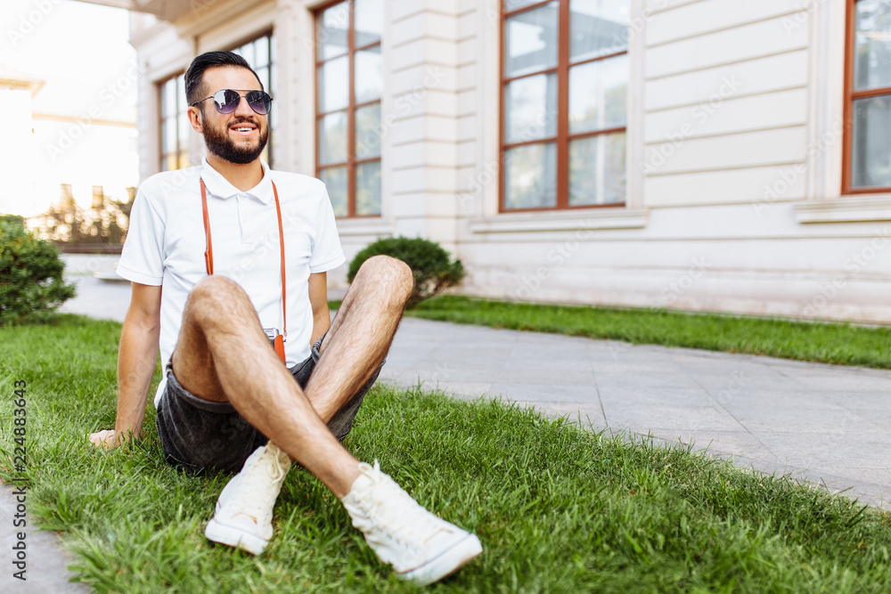 Fototapeta premium Stylish hipster with a camera on his neck, sitting on the lawn in the Park