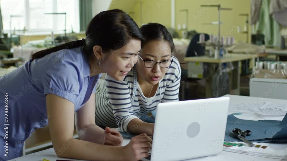 Two interested Asian women using laptop computer at sewing factory, discussing work and smiling Two interested Asian women using laptop computer at sewing factory, discussing work and smiling