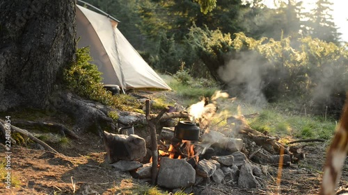 Happy adventurer drinks coffee and meditates next to the bonfire on a green mountain meadow next to tent - stunning morning during travel in the mountains.