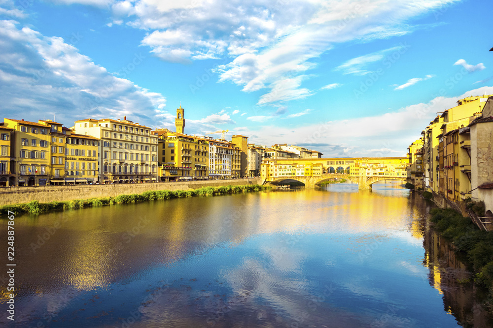 Panoramic view of the Arno River and stone medieval bridge Ponte Vecchio with beautiful reflection of colorful houses and blue sky with porous clouds, Florence, Tuscany, Italy.