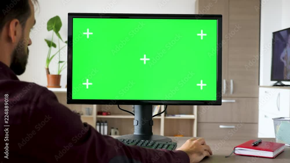 Side view of man sitting in front of computer with big green screen mock-up chroma typing on the keyboard. He is in the living room of his house