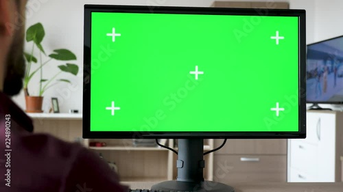 Unrecognisable man looking at modern personal computer with big green screen chroma mock up while the TV in the living room is on