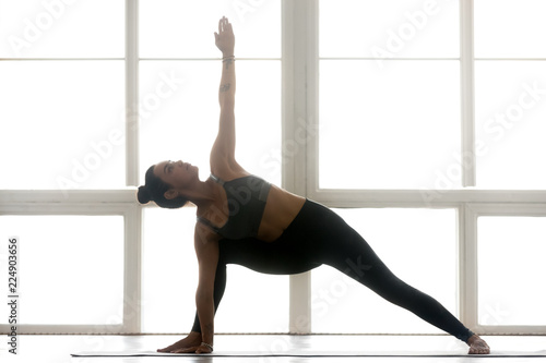 Young sporty attractive woman practicing yoga, doing Utthita parsvakonasana pose, Extended Side Angle exercise, working out, wearing sportswear, grey pants, top, indoor full length, at yoga studio