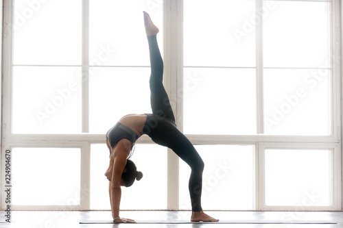 Young sporty attractive woman practicing yoga, doing Bridge exercise, One legged Wheel pose, working out, wearing sportswear, grey pants, top, indoor full length, at yoga studio, side view