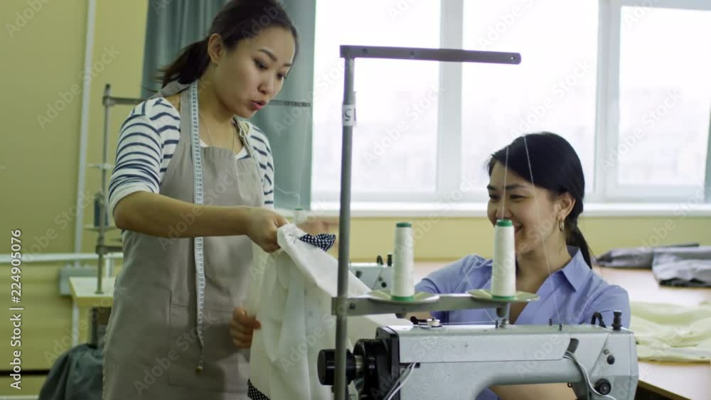 Tilt up shot of two smiling Asian women examining new shirt and talking while working at sewing factory