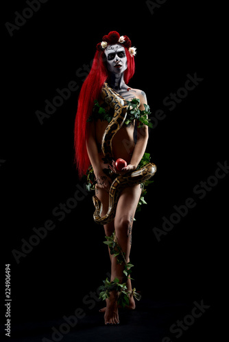 Nude scary zombie woman with red hair holding long snake piton around neck and big red apple.