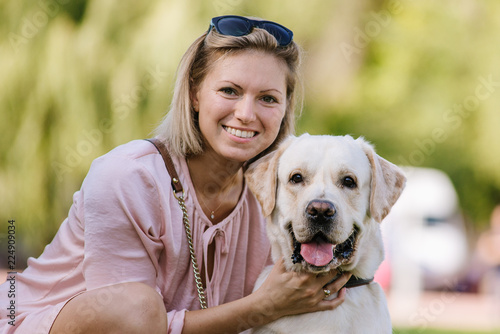 A beautiful blonde is sitting near her labrador retriever on the grass in the park.