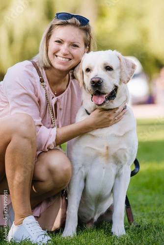 A beautiful blonde is sitting near her labrador retriever on the grass in the park.