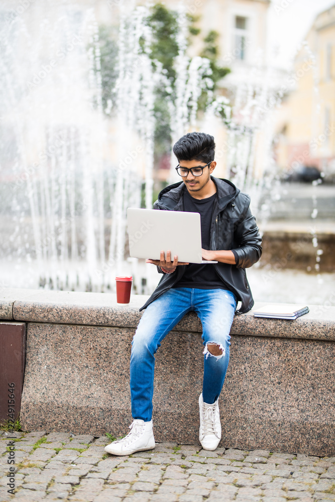 Obraz premium Handsome indian man with laptop while sitting near the fountain in the city center on a sunny summer day