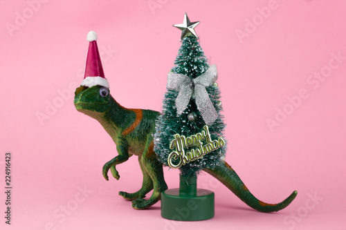 funny green dinosaur toy in little santa claus hat  near little christmas tree  on pastel pink background © dvulikaia