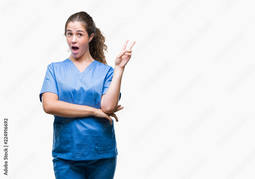 Young brunette doctor girl wearing nurse or surgeon uniform over isolated background smiling with happy face winking at the camera doing victory sign. Number two. Young brunette doctor girl wearing nurse or surgeon uniform over isolated background smiling with happy face winking at the camera doing victory sign. Number two.