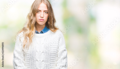 Beautiful young blonde woman wearing winter sweater over isolated background with serious expression on face. Simple and natural looking at the camera.