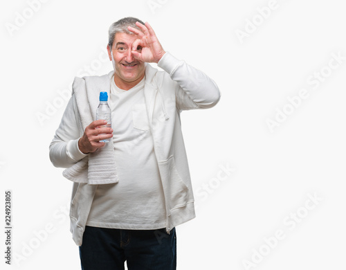 Handsome senior man training holding towel and water bottle over isolated background with happy face smiling doing ok sign with hand on eye looking through fingers