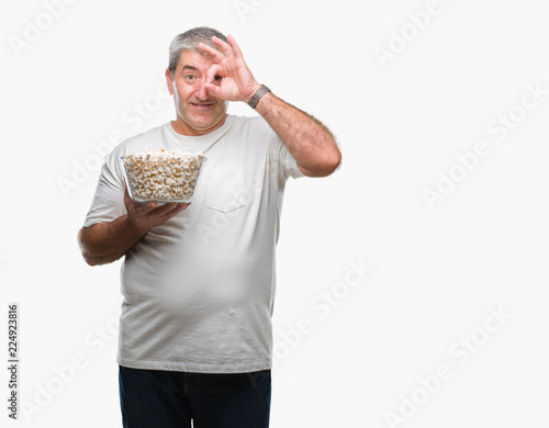 Handsome senior man eating popcorn over isolated background with happy face smiling doing ok sign with hand on eye looking through fingers
