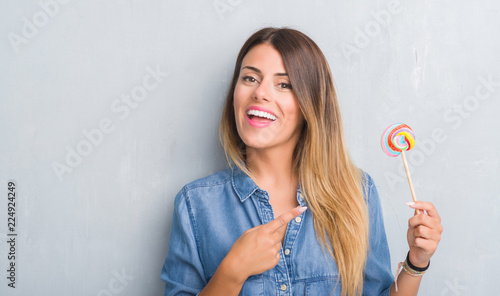 Young adult woman over grey grunge wall eating lollipop candy very happy pointing with hand and finger